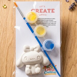 Könnte beinhalten: Ein Mal-dich-selbst-Set mit einer weißen Keramikfigur, Farbtöpfen in Gelb und Blau und einem Pinsel. Das Set ist in einer transparenten Plastiktüte mit dem Text "LET'S CREATE MAGIC!" und "Cottage Creations" verpackt.