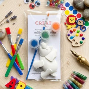 Könnte beinhalten: Ein Bastelset mit einer weißen Elefantenfigur, Farbtöpfen, Pinseln, Wackelaugen, Glitzerröhrchen und einer Holzpalette. Das Set enthält das Wort "ART" in bunten Buchstaben und den Text "LET CREATE MAGIC!"