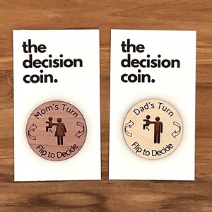 May include: Two wooden coins with the text "the decision coin." One coin has a woman holding a baby and the text "Mom's Turn" and "Flip to Decide". The other coin has a man holding a baby and the text "Dad's Turn" and "Flip to Decide".