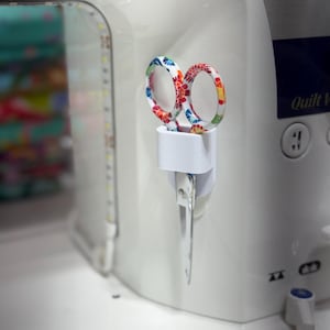 May include: A white sewing machine with a pair of colorful scissors in a holder. The scissors have floral patterns in red, blue, and yellow. The machine has Japanese text and the words "Quilt V" on it.