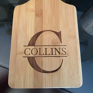 May include: A natural wood cutting board with a handle. The board is engraved with the letter "C" and the name "COLLINS" underneath.
