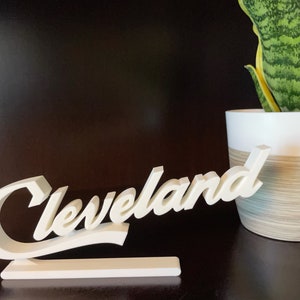May include: White 3D sign with the word "Cleveland" in a cursive font. The sign is on a white base and is sitting on a dark brown surface.