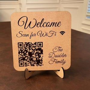 A light brown wooden sign with "Welcome Scan for WiFi" and a WiFi symbol at the top, a QR code in the middle, and "The Doubler Family" at the bottom, all in dark brown text.