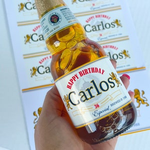 May include: A clear glass bottle of beer with a gold label. The label has the text "Happy Birthday Carlos" and "Especial 36" printed on it. The bottle is being held by a hand.