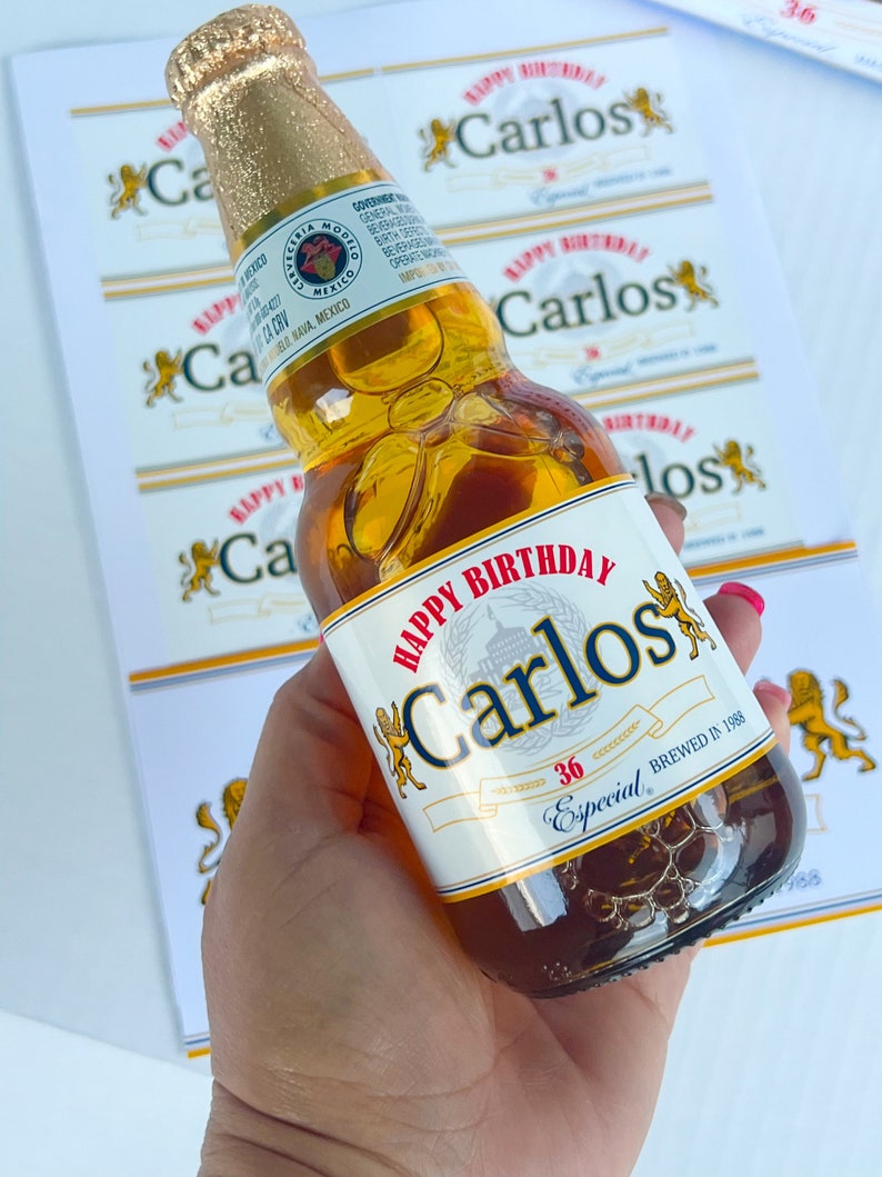 May include: A clear glass bottle of beer with a gold label. The label has the text "Happy Birthday Carlos" and "Especial 36" printed on it. The bottle is being held by a hand.