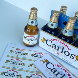 May include: A six-pack of beer with personalised labels that say "Happy Birthday Carlos" in red and gold lettering. The labels also feature a gold lion graphic. The beer bottles are in a blue and white cardboard box.