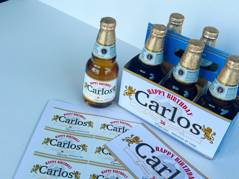 May include: A six-pack of beer with personalised labels that say "Happy Birthday Carlos" in red and gold lettering. The labels also feature a gold lion graphic. The beer bottles are in a blue and white cardboard box.