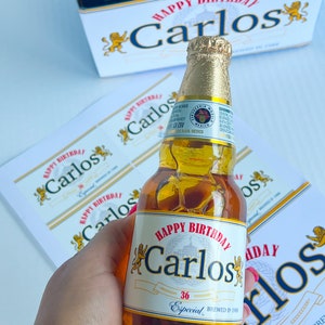 May include: A personalised beer bottle with a white label that says "Happy Birthday Carlos" with a lion graphic on either side of the text. The label also says "36 Especial Brewed in 1988".