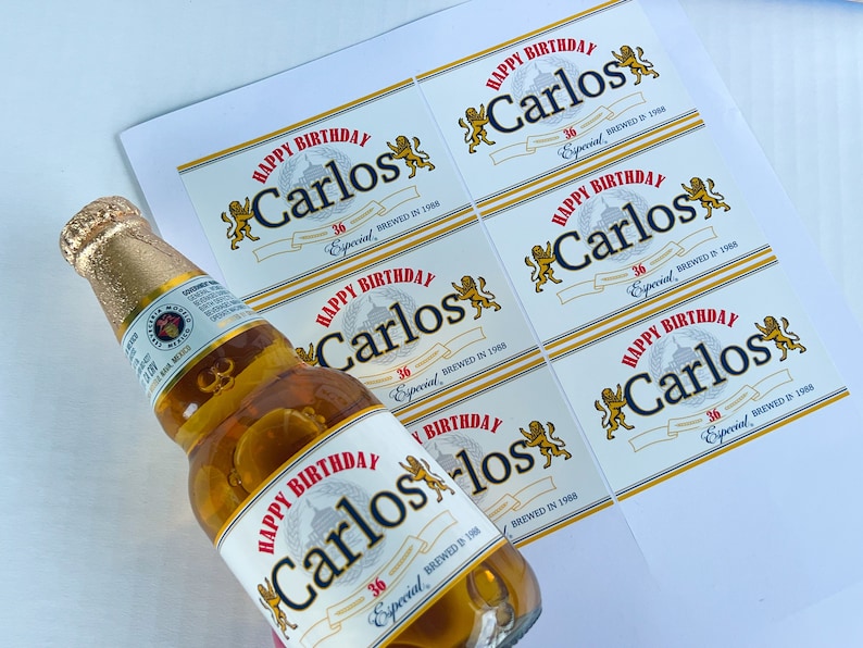 May include: A set of six white labels with a gold border and a red and gold design. The labels feature the text "Happy Birthday Carlos" with the year "1988" and the words "36 Especial" printed on them. The labels are designed to be placed on beer bottles.