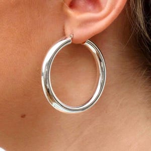 Puede incluir: Pendientes de aro de plata con una banda gruesa y redondeada. Los pendientes son grandes y tienen un diseño simple y clásico.