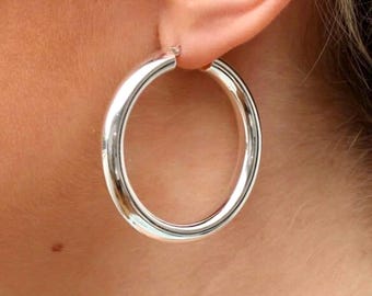 May include: Silver hoop earrings with a thick, rounded band. The earrings are large and have a simple, classic design.