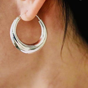 May include: Silver hoop earrings with a thick, rounded design. The earrings are large and have a smooth, polished finish.
