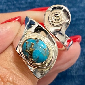 May include: A silver ring featuring a turquoise stone set within a geometric design. The ring has a distinctive, abstract shape with a spiral detail on one side.
