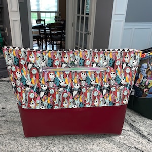May include: A large tote bag with a red bottom and a black and white patterned top. The pattern features characters from the Nightmare Before Christmas movie. The bag has a zipper closure.