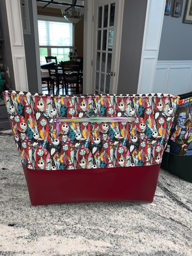May include: A large tote bag with a red bottom and a black and white patterned top. The pattern features characters from the Nightmare Before Christmas movie. The bag has a zipper closure.