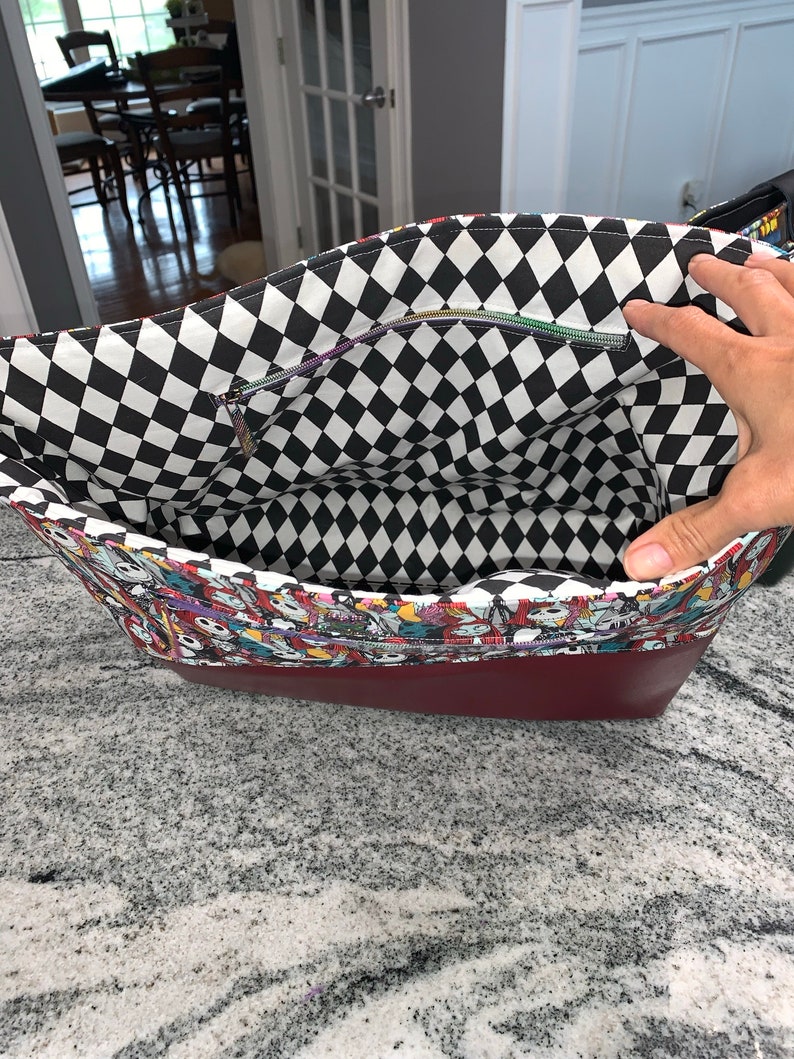 May include: A red and black checkered fabric tote bag with a zippered pocket. The bag is lined with a colorful fabric featuring characters from the Nightmare Before Christmas.