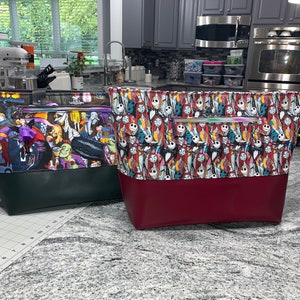 May include: Two large tote bags with zippered pockets. The bag on the left has a green bottom and a colorful print of characters from the movie "The Nightmare Before Christmas". The bag on the right has a burgundy bottom and a colorful print of characters from the movie "The Nightmare Before Christmas".