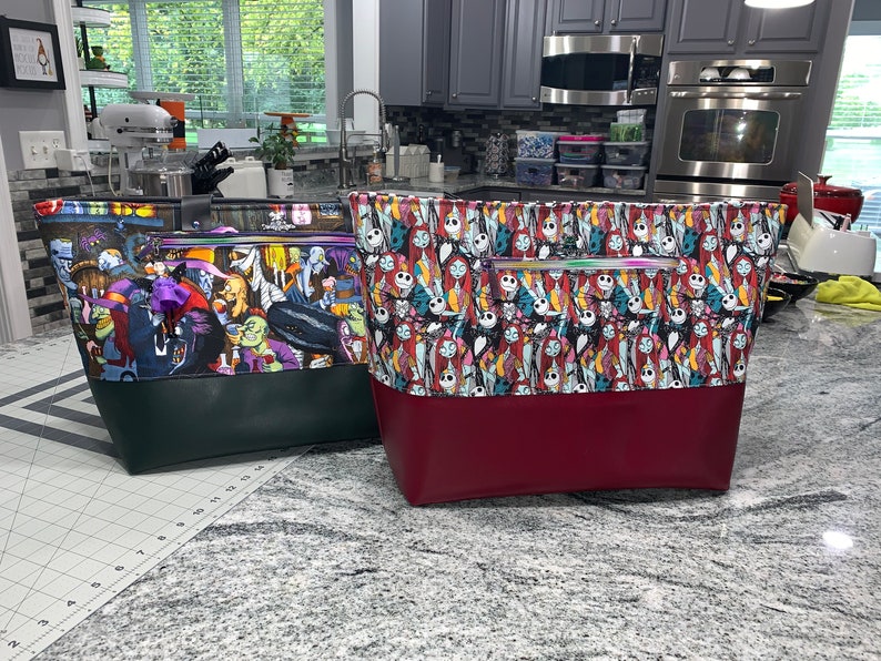 May include: Two large tote bags with zippered pockets. The bag on the left has a green bottom and a colorful print of characters from the movie "The Nightmare Before Christmas". The bag on the right has a burgundy bottom and a colorful print of characters from the movie "The Nightmare Before Christmas".