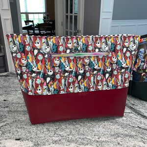 May include: A large zippered tote bag with a red bottom and a white and black patterned top featuring characters from the Nightmare Before Christmas. The pattern includes Jack Skellington, Sally, and other characters from the film.