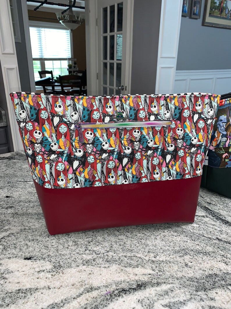 May include: A large zippered tote bag with a red bottom and a white and black patterned top featuring characters from the Nightmare Before Christmas. The pattern includes Jack Skellington, Sally, and other characters from the film.