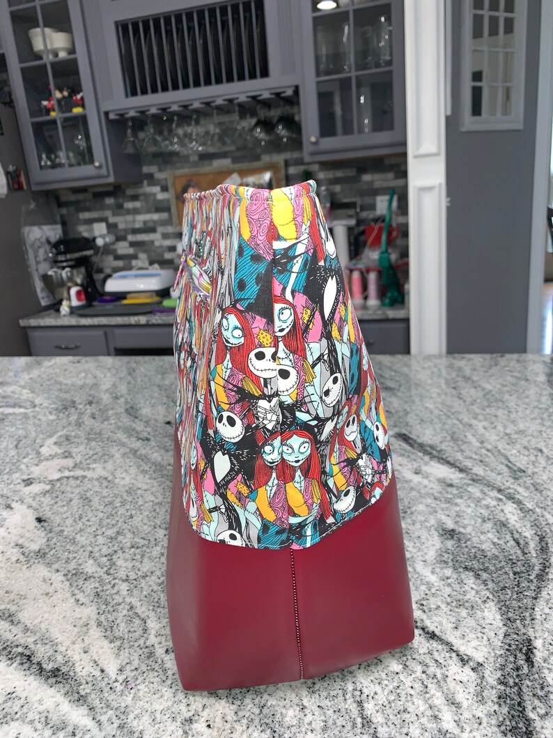 May include: A red tote bag with a colorful print of characters from The Nightmare Before Christmas. The print features Jack Skellington, Sally, and other characters from the film.