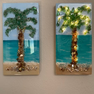 May include: Two rectangular glass art pieces depicting palm trees on a beach scene. The palm trees are made of green glass pieces and the trunks are made of brown glass pieces. The beach scene is made of blue, white, and brown glass pieces. The art pieces are lit from behind, creating a warm glow.