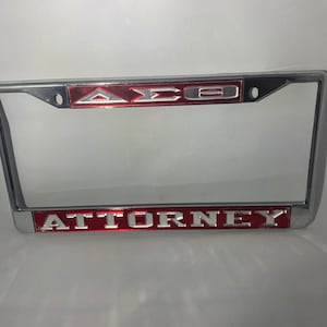 May include: A silver license plate frame with a red background and the letters "ATTORNEY" in silver lettering. The top of the frame has a silver Greek letter Delta, Sigma, Theta in red lettering.