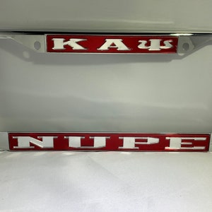May include: A silver license plate frame with red and white lettering. The top line reads "KAΨ" and the bottom line reads "NUPE".
