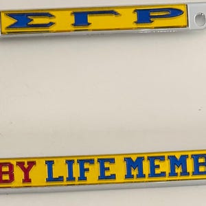 May include: A silver license plate frame with yellow and blue lettering. The top bar reads "ΣΓΡ" and the bottom bar reads "RUBY LIFE MEMBER". The frame is rectangular with rounded corners.