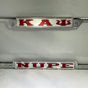 May include: A silver license plate frame with two red and white inserts. The top insert reads "KAΨ" and the bottom insert reads "NUPÉ".