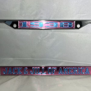 May include: A chrome license plate frame with a pink and blue background. The frame has the number "1938" and the text "JACK & JILL OF AMERICA" on it.
