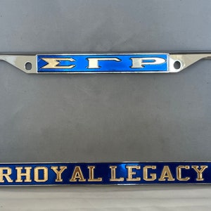May include: A chrome license plate frame with a blue and gold design. The top of the frame says "ΣΓΡ" and the bottom says "RHOyal LEGACY".