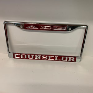 May include: A chrome license plate frame with red lettering that says "AXE" and "COUNSELOR".