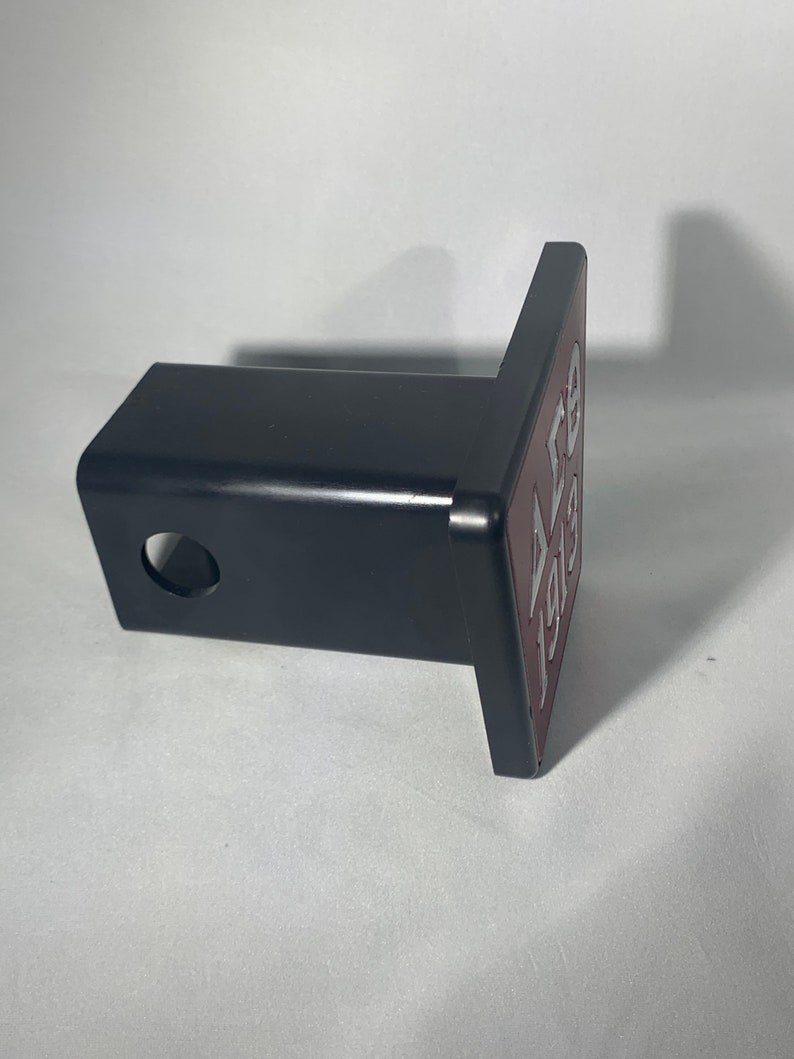 Puede incluir: Un receptor de enganche de metal negro con una abertura cuadrada y un agujero redondo. El receptor tiene una placa de color burdeos unida a &eacute;l con el texto "ALB 193" en letras plateadas.