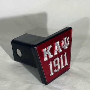 May include: Black trailer hitch cover with a red and silver emblem. The emblem features the Greek letters KAU and the number 1911. The hitch cover is made of metal and has a hole for a pin.