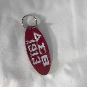 May include: A red oval-shaped keychain with a silver metal ring. The keychain has white lettering that reads "BMA 1913" with a triangle symbol.