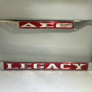 May include: A silver license plate frame with a red background and white lettering. The top of the frame says "ΔΣΘ" and the bottom says "LEGACY".