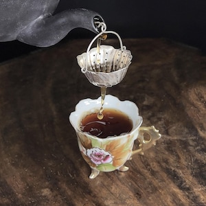 May include: A silver tea strainer with a handle is suspended from a dark teapot spout, filtering liquid into a small porcelain teacup. The teacup features a painted pink rose, green leaves, and gold trim, resting on three small gold-coloured feet.