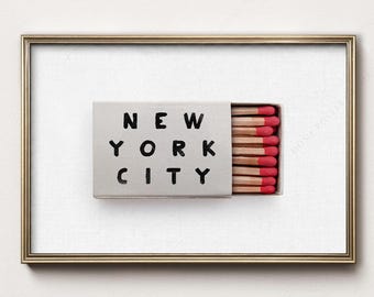 May include: A framed print featuring a light gray matchbox with the text NEW YORK CITY printed in black capital letters. The matchbox is partially open, revealing a row of wooden matches with bright red tips inside the box.