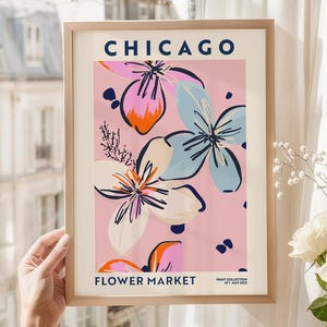 May include: A framed wall art print titled CHICAGO FLOWER MARKET with a pink background featuring stylized floral illustrations in shades of blue, orange, and cream. The text PRINT COLLECTION N&deg;1 JULY 2022 is printed in the bottom right corner.