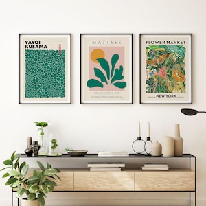 May include: Three framed art prints hanging on a wall above a wooden console table. The left print features green dots by Yayoi Kusama. The center print shows a Matisse abstract design. The right print displays a colorful Flower Market New York graphic.