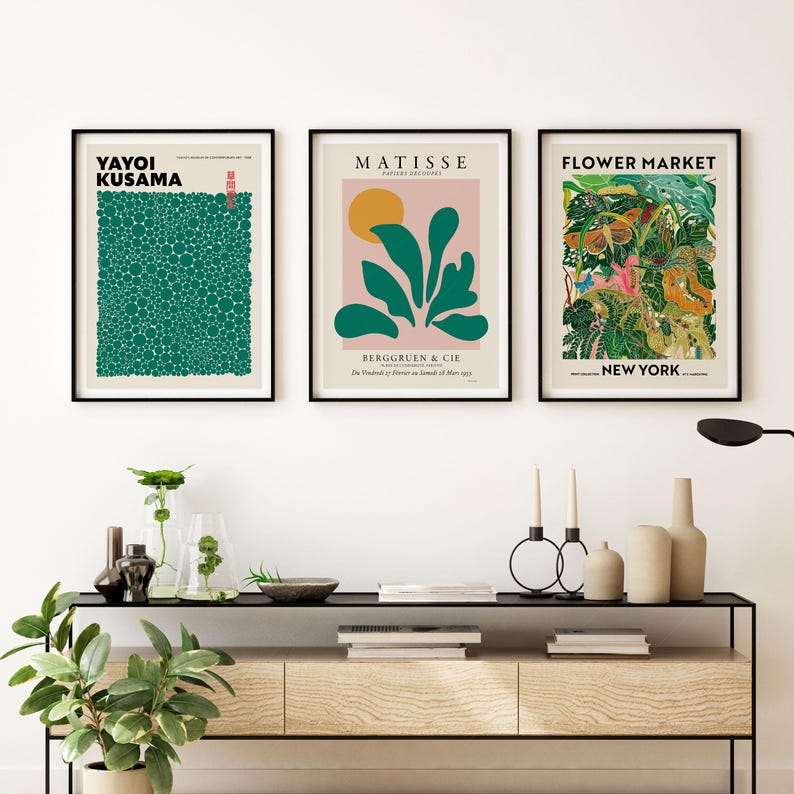 May include: Three framed art prints hanging on a wall above a wooden console table. The left print features green dots by Yayoi Kusama. The center print shows a Matisse abstract design. The right print displays a colorful Flower Market New York graphic.