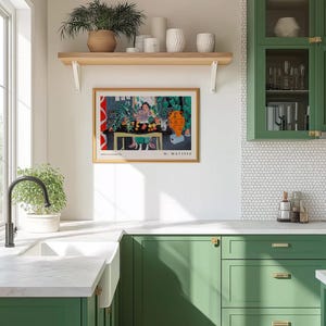 May include: A framed Henri Matisse art print featuring a colorful interior scene with a person at a table, displayed on a white wall above green kitchen cabinets, a white farmhouse sink, and a wooden shelf holding various white ceramic vases.