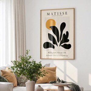 May include: A framed wall art print titled MATISSE PAPIERS D&Eacute;COUPES featuring a large yellow circle and black organic leaf-like shapes on a light beige background, with the text BERGGRUEN & CIE and exhibition dates printed at the bottom.