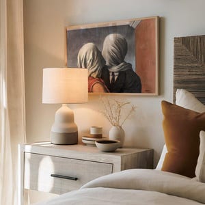 May include: A framed print of Rene Magritte's The Lovers, featuring two figures with heads covered in white fabric, hanging above a bedside table. The table holds a two-toned ceramic lamp, a small vase with dried stems, and stacked ceramic bowls.