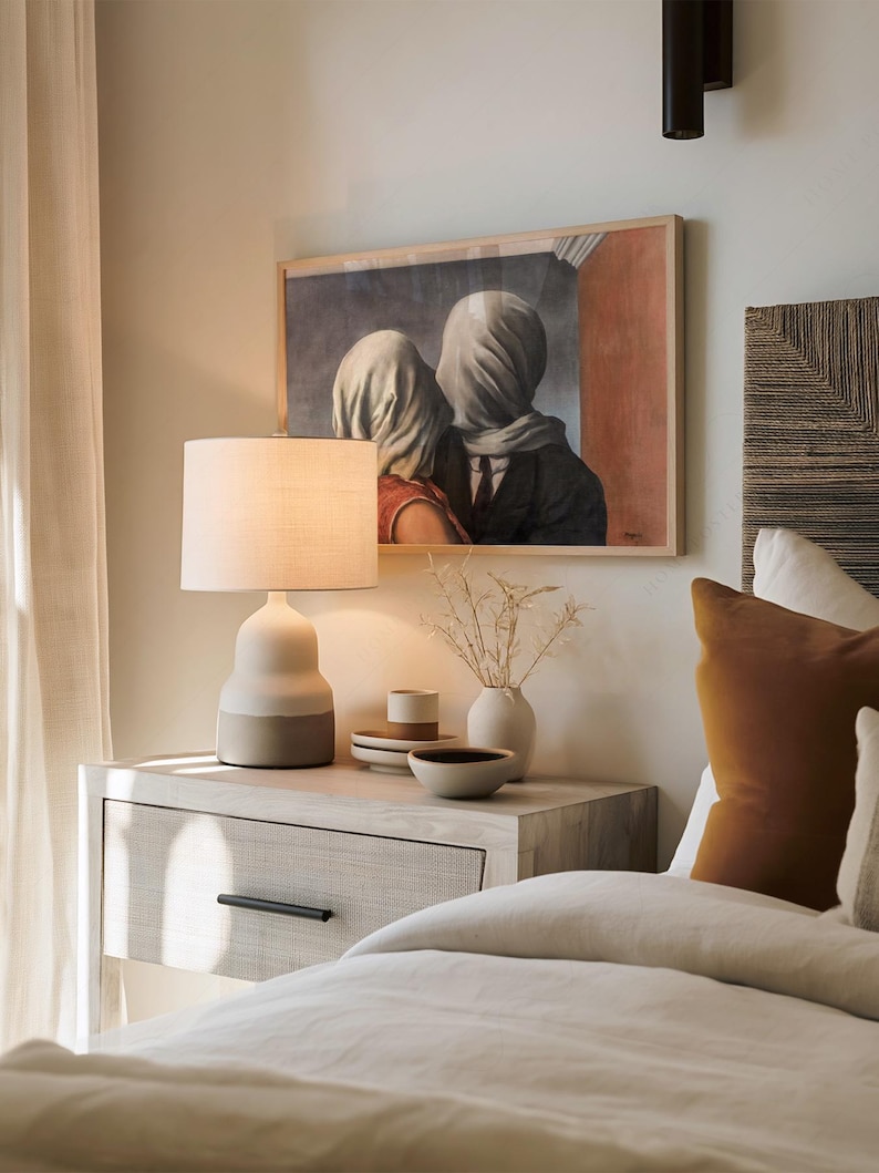 May include: A framed print of Rene Magritte's The Lovers, featuring two figures with heads covered in white fabric, hanging above a bedside table. The table holds a two-toned ceramic lamp, a small vase with dried stems, and stacked ceramic bowls.