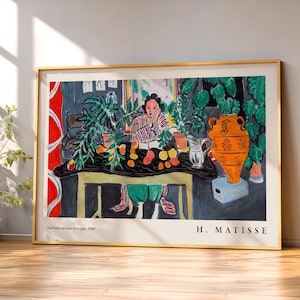 May include: A framed art print of Henri Matisse's painting Int&eacute;rieur au vase &eacute;trusque, 1940. The artwork depicts a person sitting at a table with a large orange vase, green plants, and fruit, rendered in a colorful, expressive style with bold lines.