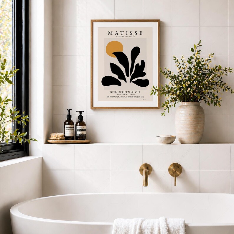 May include: A framed Matisse Papiers D&eacute;coup&eacute;s exhibition poster featuring a black abstract plant shape and a yellow circle on a light beige background, displayed on a white tiled wall above a bathtub with two dark brown pump bottles.