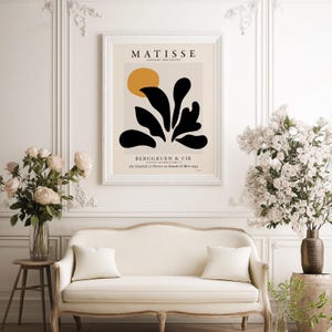May include: A framed wall art print featuring a Matisse Papiers D&eacute;coup&eacute;s exhibition poster. The design includes a mustard yellow circle above abstract black organic shapes on a beige background, with text reading MATISSE, BERGGRUEN & CIE.