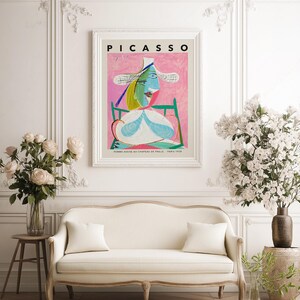 May include: A framed art print titled PICASSO featuring the painting Femme assise au chapeau de paille. The artwork displays a stylized figure in a wide-brimmed hat with blue, green, and yellow tones against a textured pink background.